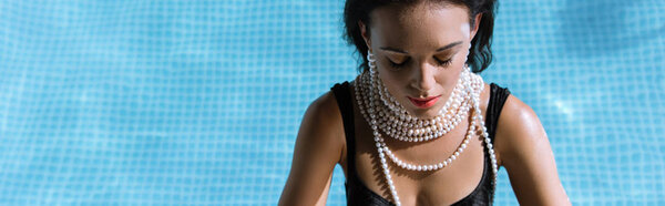 panoramic shot of attractive woman in black swimsuit and pearl necklace posing in pool 