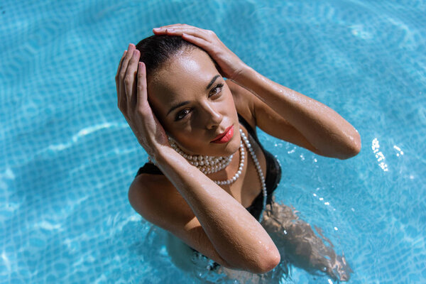 high angle view of attractive woman in black swimsuit and pearl necklace posing in pool 
