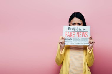 sad asian woman in yellow outfit holding newspaper with fake news isolated on pink