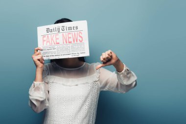 woman in white blouse holding newspaper with fake news and showing thumb down on blue background clipart