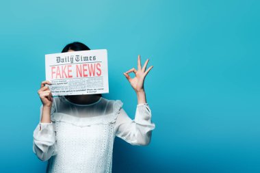 woman in white blouse holding newspaper with fake news and showing ok sign on blue background clipart