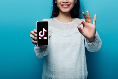 KYIV, UKRAINE - JULY 15, 2019: cropped view of smiling asian woman in white blouse showing ok sign and smartphone with tiktok app on blue background clipart