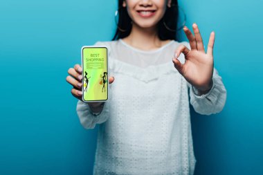 Beyaz bluzlu gülümseyen Asyalı kadın mavi arka planda en iyi alışveriş uygulamasına sahip işaret ve akıllı telefon gösteriyor.