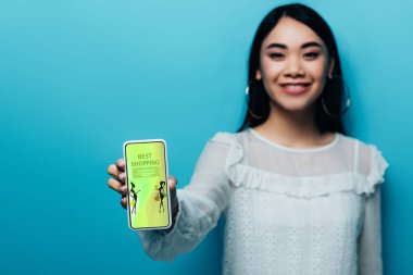 Beyaz bluzlu gülümseyen Asyalı kadının elinde mavi arka planda moda alışverişi uygulaması olan akıllı telefon tutuşu.