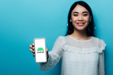 Kyiv, Ukrayna - 15 Temmuz 2019: Beyaz bluzlu gülümseyen Asyalı kadının seçici odak noktası mavi arka planda android logosu olan akıllı telefon tutması.