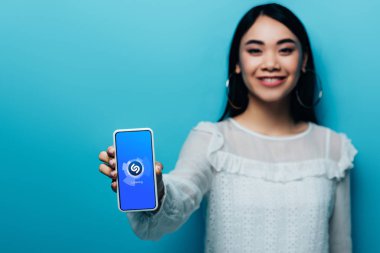 Kyiv, Ukrayna - 15 Temmuz 2019: Beyaz bluzlu gülümseyen Asyalı kadının seçici odak noktası mavi arka planda shazam uygulamalı akıllı telefon tutması.