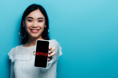 Kyiv, Ukrayna - 15 Temmuz 2019: Beyaz bluzlu gülümseyen Asyalı kadının seçici odak noktası mavi arka planda netflix uygulamalı akıllı telefon tutması.