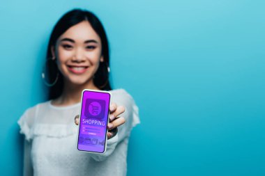 Beyaz bluzlu gülümseyen Asyalı kadının elinde mavi arka planda online alışveriş uygulaması olan akıllı telefon tutuşu.