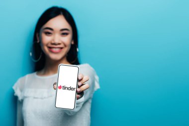 Kyiv, Ukrayna - 15 Temmuz 2019: Beyaz bluzlu gülümseyen Asyalı kadının seçici odak noktası mavi arka planda Tinder uygulamalı akıllı telefon tutması