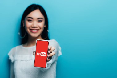 Kyiv, Ukrayna - 15 Temmuz 2019: Beyaz bluzlu gülümseyen Asyalı kadının seçici odak noktası mavi arka planda youtube uygulaması ile akıllı telefon tutması