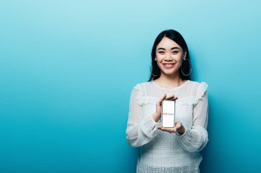 KYIV, UKRAINE - JULY 15, 2019: smiling asian woman in white blouse holding smartphone with gmail app on blue background clipart