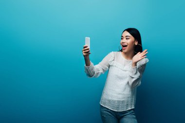excited asian woman in white blouse having video chat on smartphone and waving hand on blue background clipart