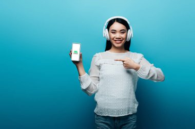 Kyiv, Ukrayna - 15 Temmuz 2019: kulaklıklı gülümseyen Asyalı kadın mavi arka planda Android simgesi olan akıllı telefonu işaret ediyor