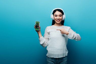 Kulaklıklı gülümseyen Asyalı kadın mavi arka planda kayıt uygulaması olan akıllı telefonu işaret ediyor.