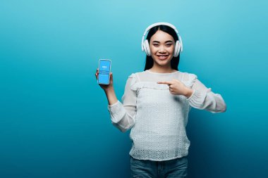 Kyiv, Ukrayna - 15 Temmuz 2019: kulaklıklı gülümseyen Asyalı kadın mavi arka planda skype simgesi olan akıllı telefonu işaret ediyor