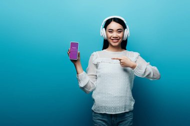 Kyiv, Ukrayna - 15 Temmuz 2019: Mavi arka planda Instagram uygulaması olan akıllı telefonu işaret eden kulaklıklı gülümseyen Asyalı kadın