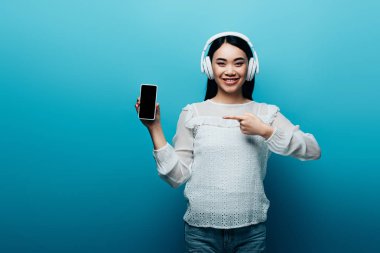 Kulaklıklı gülümseyen Asyalı kadın mavi arka planda boş ekranlı akıllı telefonu işaret ediyor.