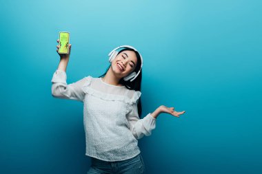 Yüzünde kulaklıkla gülümseyen Asyalı kadın elinde en iyi alışveriş uygulaması olan akıllı telefon ve mavi arka planda dans eden.