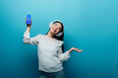 Kyiv, Ukrayna - 15 Temmuz 2019: kulaklıkları kapalı gülümseyen Asyalı kadın Shazam uygulamalı akıllı telefon tutuyor ve mavi arka planda dans ediyor.