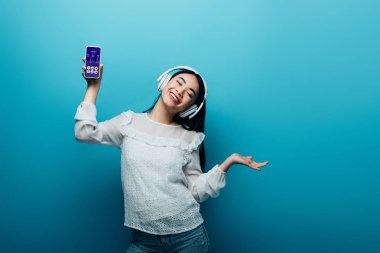 Yüzünde kulaklıkla gülümseyen Asyalı kadın. Elinde sağlık uygulaması olan akıllı telefon, mavi arka planda dans ediyor.