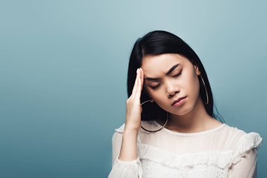 upset brunette asian woman with headache on blue background clipart