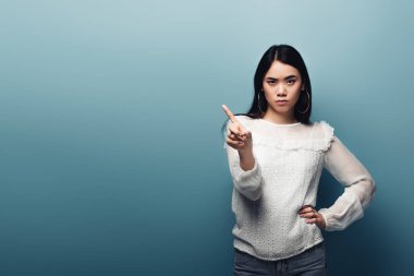 upset brunette asian woman with hand on hip showing no gesture on blue background clipart