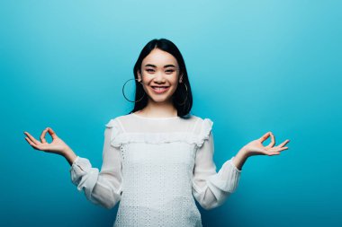 happy brunette asian woman meditating on blue background clipart