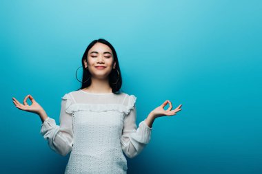 Mutlu esmer Asyalı kadın, mavi arka planda gözleri kapalı meditasyon yapıyor.