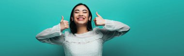happy brunette asian woman showing thumbs up on turquoise background, panoramic shot clipart