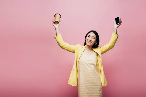 黄色の衣装を着た幸せなアジア人女性が紙コップとスマートフォンをピンクに隔離し — ストック写真