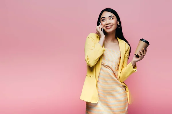 黄色の衣装を着た幸せなアジア人女性が紙コップを持ってピンクに隔離されたスマートフォンで話しています — ストック写真