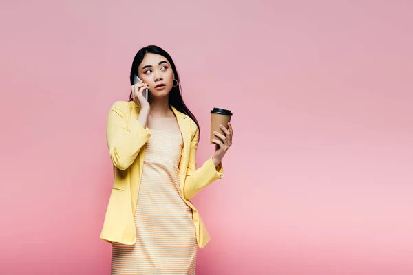 黄色の衣装を着たアジア人女性が紙コップを持ってピンクに隔離されたスマートフォンで話しています — ストック写真