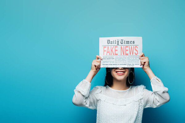 smiling asian woman in white blouse holding newspaper with fake news on blue background