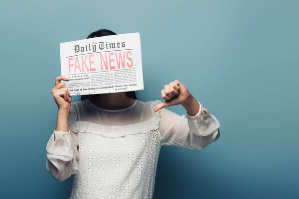 woman in white blouse holding newspaper with fake news and showing thumb down on blue background