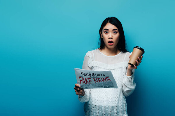 shocked asian woman in white blouse holding newspaper with fake news and paper cup on blue background