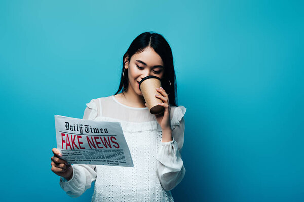 asian woman in white blouse holding newspaper with fake news and drinking coffee on blue background