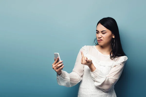 穿着白衬衫的不满意的亚洲女人在蓝色背景的智能手机屏幕上显示出中指 — 图库照片