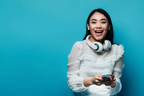 快乐的亚洲女人 穿着白衬衫 带着无线耳机 用蓝色背景的智能手机 — 图库照片