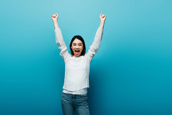 happy brunette asian woman with hands in air on blue background