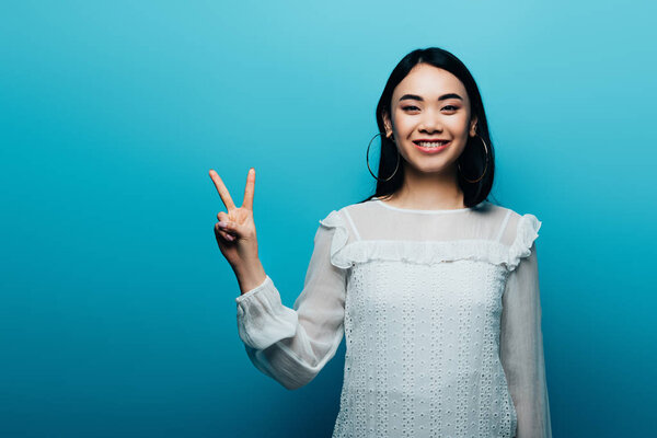 счастливая брюнетка азиатская женщина показывает знак мира на голубом фоне
