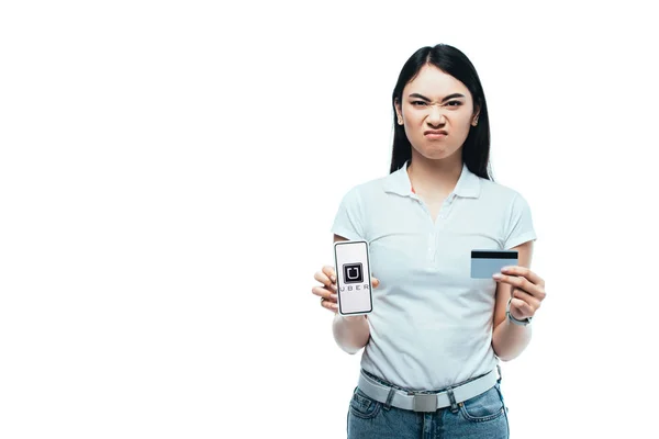stock image KYIV, UKRAINE - JULY 15, 2019: displeased brunette asian girl holding credit card and smartphone with uber app isolated on white