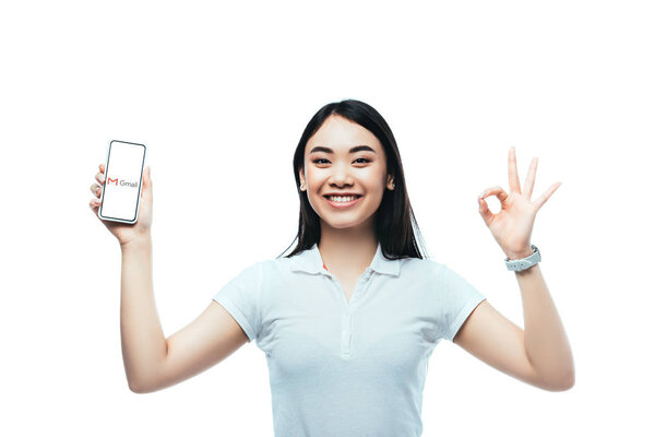 KYIV, UKRAINE - JULY 15, 2019: happy brunette asian woman holding smartphone with gmail app and showing ok sign isolated on white