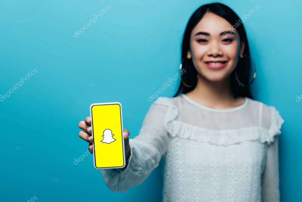 KYIV, UKRAINE - JULY 15, 2019: selective focus of smiling asian woman in white blouse holding smartphone with snapchat app on blue background