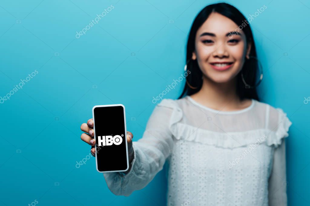 KYIV, UKRAINE - JULY 15, 2019: selective focus of smiling asian woman in white blouse holding smartphone with HBO app on blue background