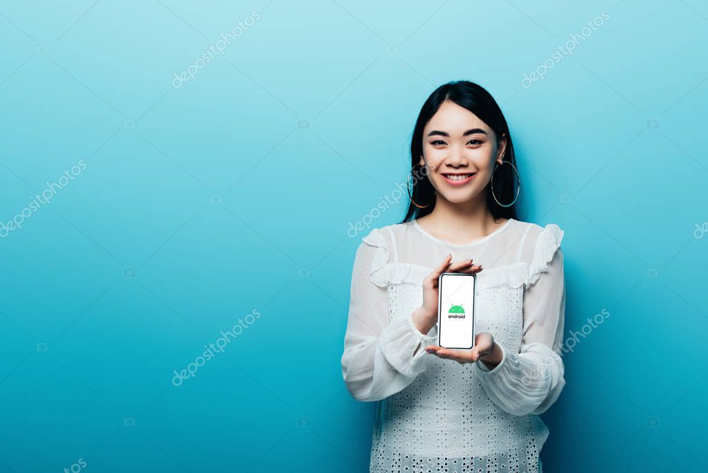 KYIV, UKRAINE - JULY 15, 2019: smiling asian woman in white blouse holding smartphone with android logo on blue background