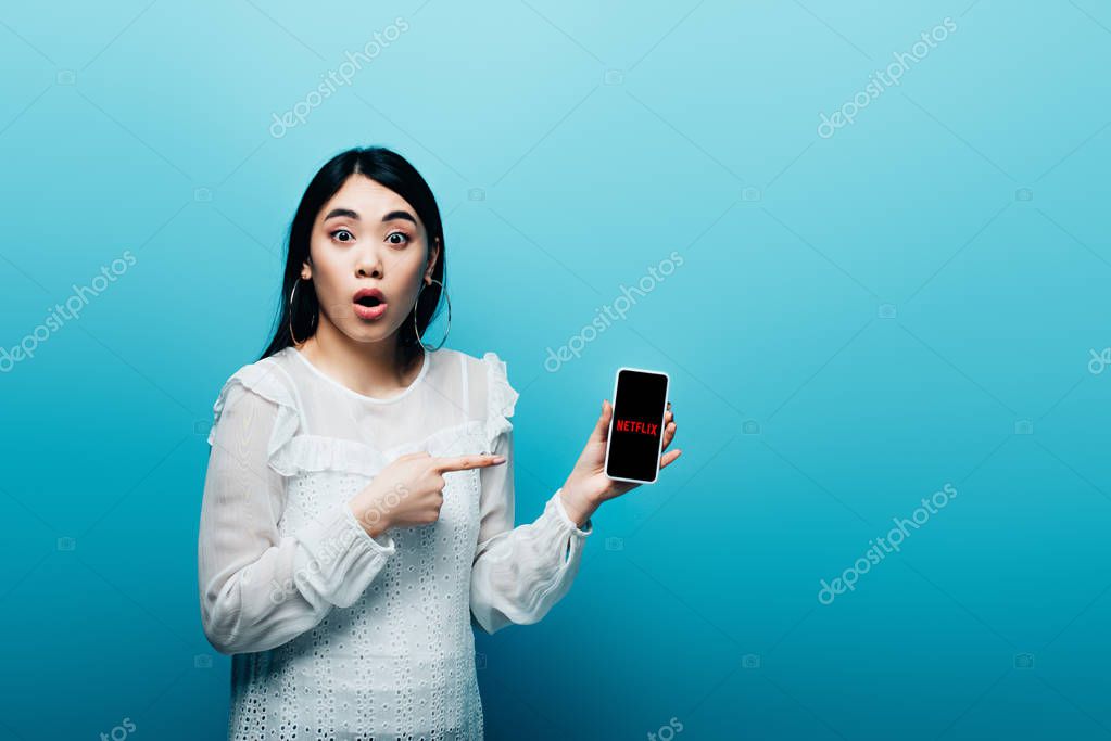 KYIV, UKRAINE - JULY 15, 2019: shocked asian woman pointing with finger at smartphone with netflix app on blue background