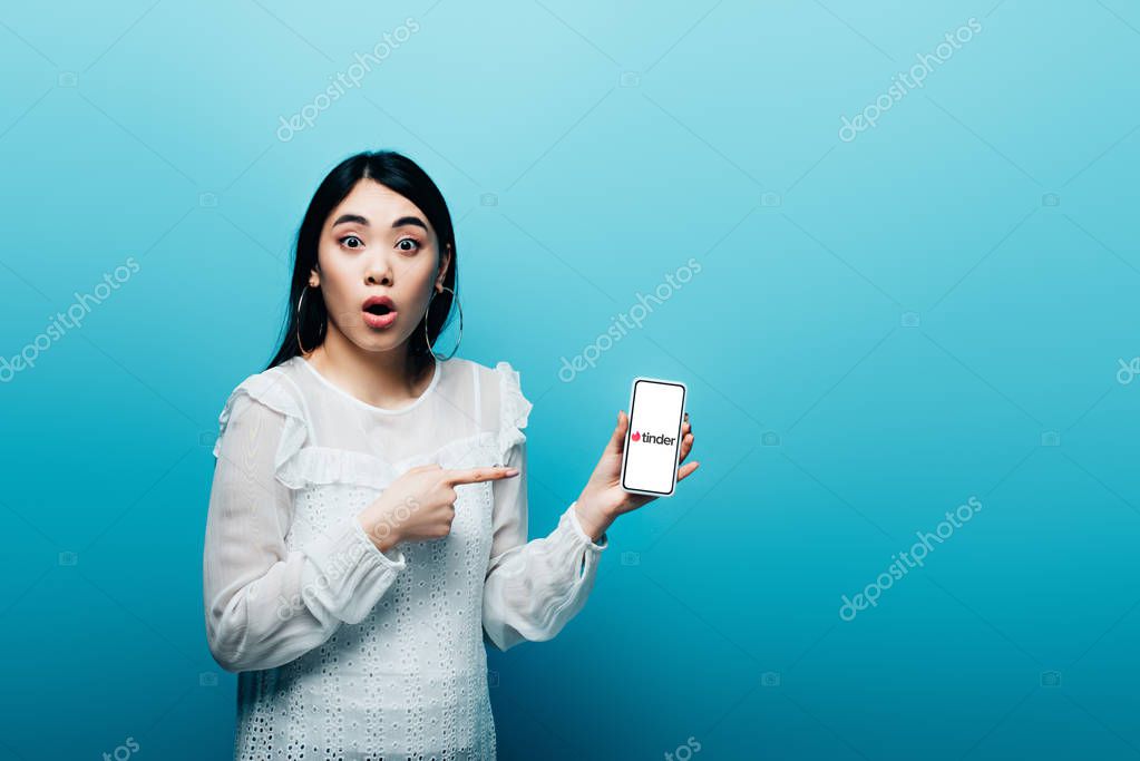 KYIV, UKRAINE - JULY 15, 2019: shocked asian woman pointing with finger at smartphone with tinder app on blue background