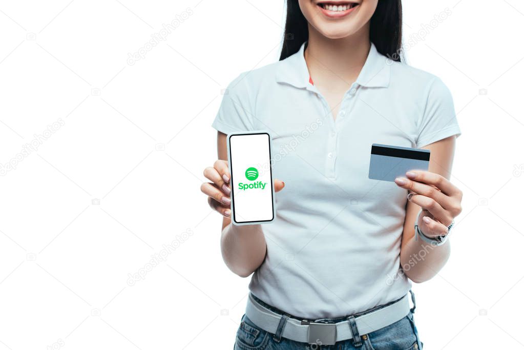 KYIV, UKRAINE - JULY 15, 2019: cropped view of smiling brunette asian girl holding credit card and smartphone with spotify app isolated on white