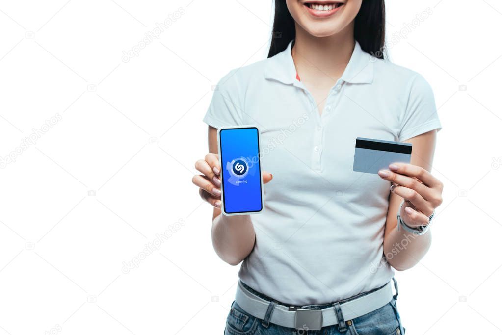 KYIV, UKRAINE - JULY 15, 2019: cropped view of smiling brunette asian girl holding credit card and smartphone with shazam app isolated on white