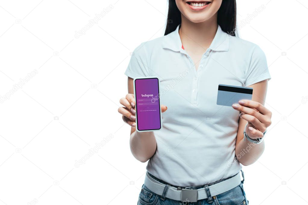 KYIV, UKRAINE - JULY 15, 2019: cropped view of smiling brunette asian girl holding credit card and smartphone with Instagram app isolated on white